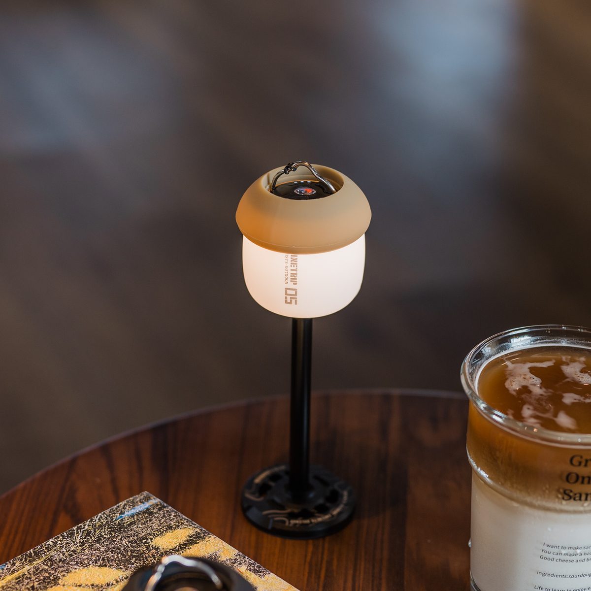 A61 Acorn Lamp with a sand shade mounted on a black light stand beside an iced drink on a wooden table